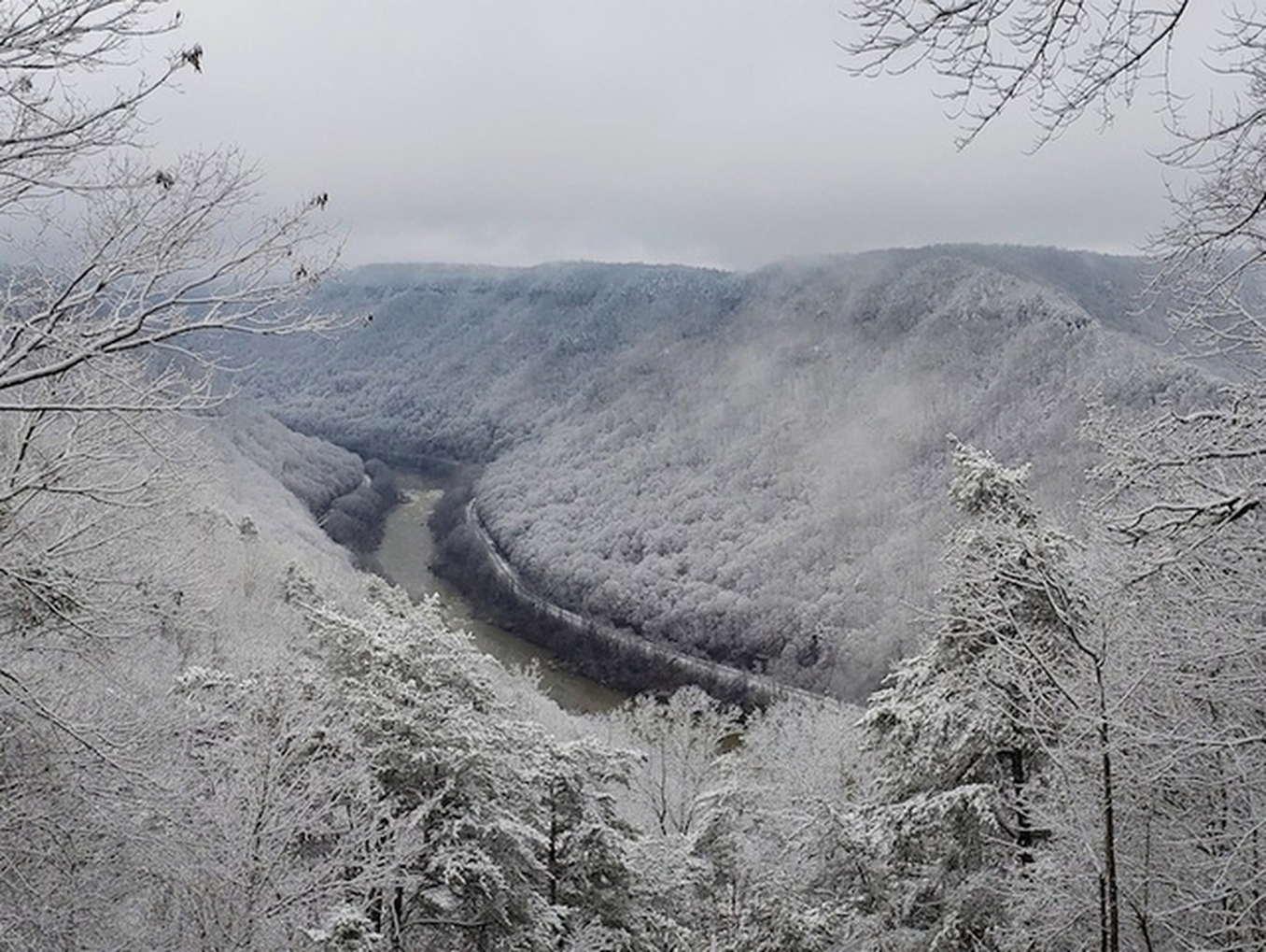 Wide scenic winter view into the New River Gorge also shows rapids below a bend and the road and railroad tracks cut into the wooded slopes on opposite sides of the river